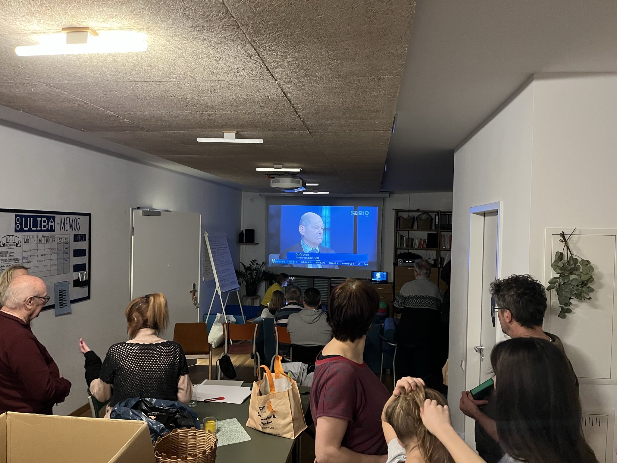 Viele Menschen im Gemeinschaftsraum verfolgen auf einer Leinwand die Wahlberichterstattung
